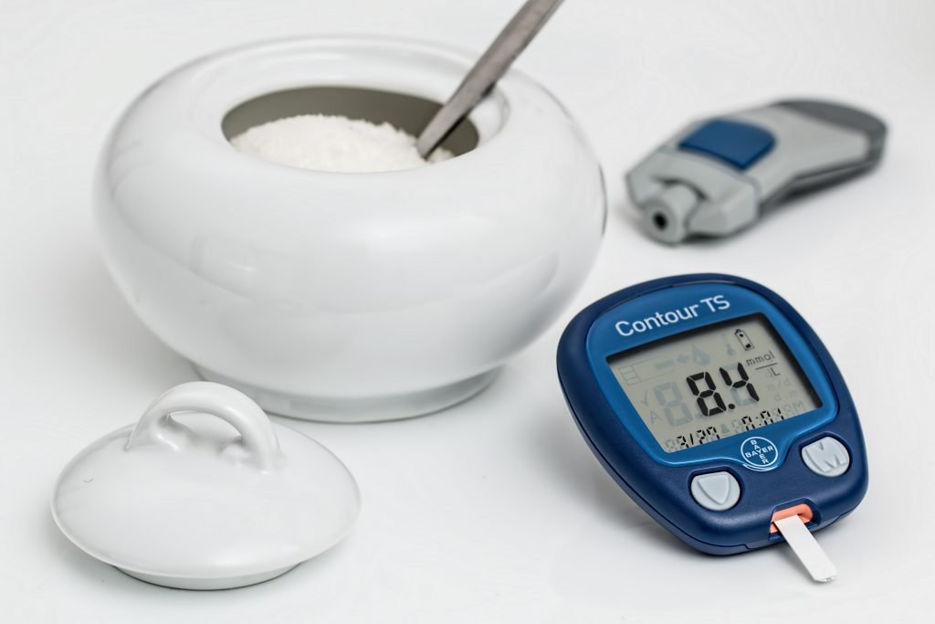 A white bowl filled with sugar with a spoon inside sitting next to a glucose monitor. Monitor reads 84.